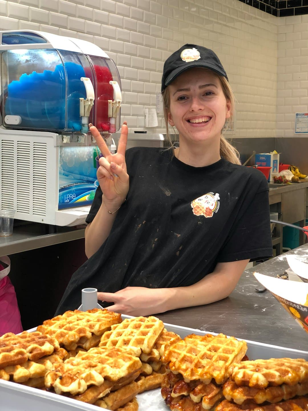 Employee in a black uniform makes a peace sign behind a waffle counter.